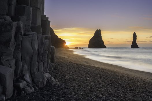 View of Reynisfjara Beach featuring basalt columns and Reynisdrangar sea stacks at sunset over the black sand shore in Reynisfjara Myrdalshreppur Iceland.