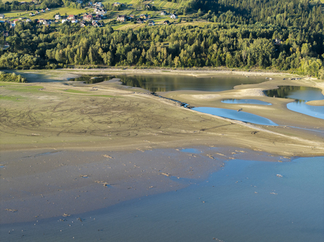 Aerial view of a reservoir with exposed mudflats and sandbanks featuring visible tire tracks on the dry lake bed surrounded by a dense forest and residential houses in Poland. Aerial view of a reservoir with exposed mudflats and sandbanks featuring visible tire tracks on the dry lake bed surrounded by a dense forest and residential houses in Poland.