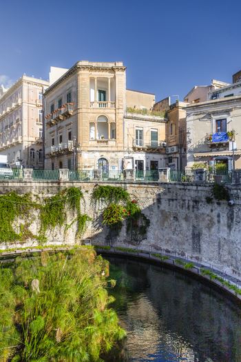 View of a serene canal with lush greenery flowing under a stone bridge shadowed by old buildings under a clear sky Ortigia Sicily Italy.