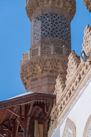 View of the Al Azhar Mosque minaret featuring intricate stone carvings and blue geometric tile patterns against a clear sky in Cairo Egypt. View of the Al Azhar Mosque minaret featuring intricate stone carvings and blue geometric tile patterns against a clear sky in Cairo Egypt.