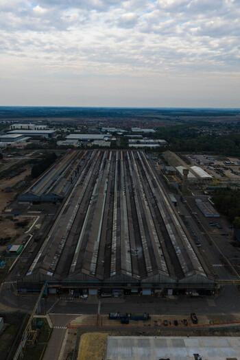 Aerial view of a large industrial factory complex with long sawtooth roofs surrounding parking areas and distant urban landscape under a cloudy sky in United Kingdom. Aerial view of a large industrial factory complex with long sawtooth roofs surrounding parking areas and distant urban landscape under a cloudy sky in United Kingdom.