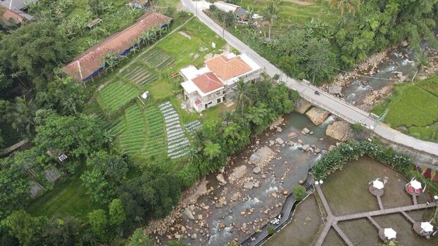 Aerial view of Kopi Tubing Cafe and Resto along a rocky river with lush rice terraces a bridge and tropical vegetation in Kecamatan Pamijahan Jawa Barat Indonesia. Aerial view of Kopi Tubing Cafe and Resto along a rocky river with lush rice terraces a bridge and tropical vegetation in Kecamatan Pamijahan Jawa Barat Indonesia.