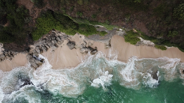 Aerial view of a sandy beach with rocky cliffs and lush vegetation meeting turquoise ocean waves in Indonesia.