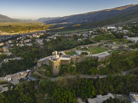 Aerial view of Gjirokaster Fortress overlooking the historic stone city with lush green hills winding roads and warm golden hour light Gjirokaster Gjirokaster County Albania. Aerial view of Gjirokaster Fortress overlooking the historic stone city with lush green hills winding roads and warm golden hour light Gjirokaster Gjirokaster County Albania.