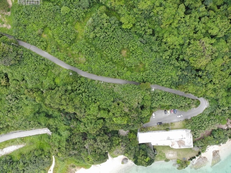 Aerial view of the Hyakume coastline featuring a winding road through dense forest a parking area and a white building near the sandy beach and emerald green sea Nanjo Okinawa Japan. Aerial view of the Hyakume coastline featuring a winding road through dense forest a parking area and a white building near the sandy beach and emerald green sea Nanjo Okinawa Japan.