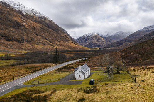 Aerial view of the iconic white cottage nestled in the dramatic Glen Coe valley with snow capped mountains and a winding road under a cloudy sky in Scotland United Kingdom. Aerial view of the iconic white cottage nestled in the dramatic Glen Coe valley with snow capped mountains and a winding road under a cloudy sky in Scotland United Kingdom.