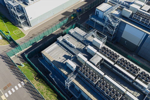 Aerial view of a data center facility with rows of industrial HVAC cooling units on the roof metal security fencing and paved grounds in the United Kingdom.