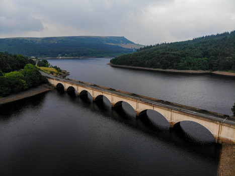 Aerial view of Ladybower Reservoir with its iconic stone arched bridge spanning the dark water surrounded by lush green hills under an overcast sky in Hope Valley England United Kingdom.