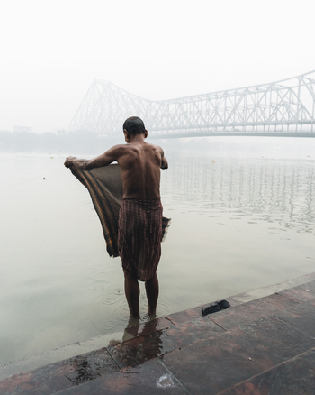 View of a man standing in the holy river Ganges drying himself with the Howrah bridge looming in the misty background Kolkata West Bengal India. View of a man standing in the holy river Ganges drying himself with the Howrah bridge looming in the misty background Kolkata West Bengal India.