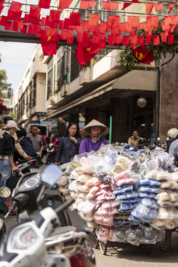 Hanoi Ha Noi Vietnam 11 November 2025 View of a street vendor in a conical hat pushing a cart filled with plastic sandals through a busy street decorated with red flags.