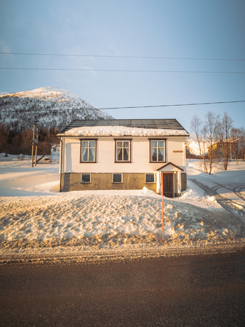 View of a quaint white house with a brown roof nestles in a snow covered landscape under a clear blue sky a mountain looms in the background Indregård Troms Norway. View of a quaint white house with a brown roof nestles in a snow covered landscape under a clear blue sky a mountain looms in the background Indregård Troms Norway.