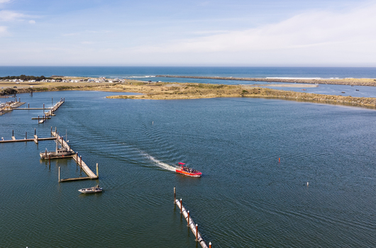Gold Beach Oregon United States 23 September 2026 Aerial view of Jerry s Rogue Jets boat navigating the Rogue River estuary with docks calm water and the Pacific Ocean in the background.
