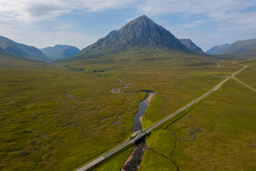Aerial view of Buachaille Etive Mor mountain rising above the green valley with a winding river flowing under a bridge and a road in Ballachulish Scotland United Kingdom. Aerial view of Buachaille Etive Mor mountain rising above the green valley with a winding river flowing under a bridge and a road in Ballachulish Scotland United Kingdom.