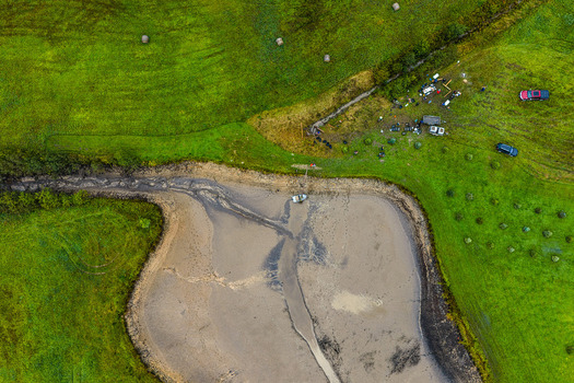 Aerial view of a muddy riverbank with a small boat surrounded by lush green fields scattered hay bales and parked vehicles in Latvia.