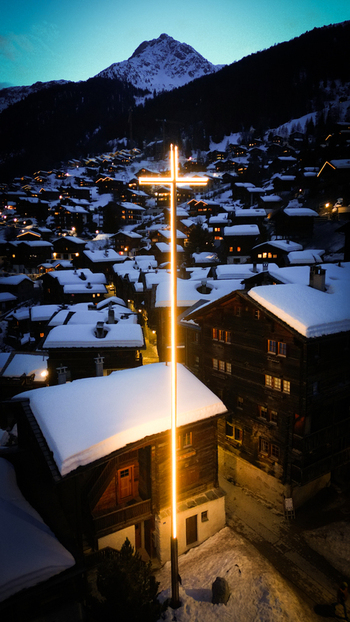Aerial view of a glowing cross piercing the night sky above snow dusted rooftops with dark wooden chalets nestled beneath a majestic snow capped mountain Grimentz Switzerland.