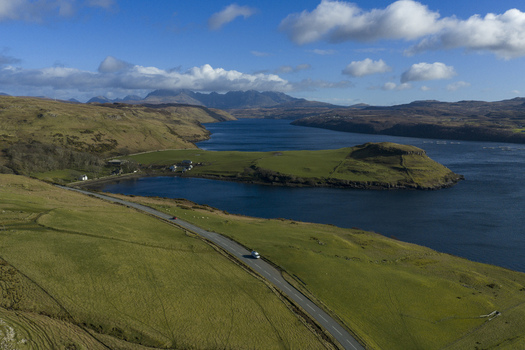 Aerial view of Gesto Bay Viewpoint featuring a winding road rolling green hills and distant mountains under a blue sky with white clouds Isle of Skye Scotland United Kingdom.