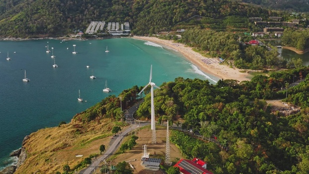 Aerial view of a wind turbine on a hill overlooking a sandy beach with boats in the turquoise sea and lush tropical vegetation in Phuket Phuket Thailand. Aerial view of a wind turbine on a hill overlooking a sandy beach with boats in the turquoise sea and lush tropical vegetation in Phuket Phuket Thailand.
