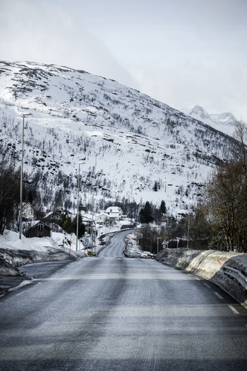 View of a winding asphalt road leading towards snow covered mountains and small houses under an overcast sky in Kvaloya Troms Norway. View of a winding asphalt road leading towards snow covered mountains and small houses under an overcast sky in Kvaloya Troms Norway.