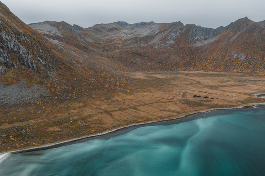 Aerial view of Senja island featuring rugged mountains with autumn foliage rocky slopes and clear turquoise coastal waters under a cloudy sky in Senja Troms Norway.