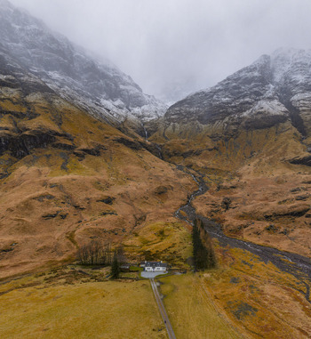 Aerial view of a small white cottage nestled at the base of dramatic snow dusted mountains and rugged valley terrain in Glencoe Scotland United Kingdom. Aerial view of a small white cottage nestled at the base of dramatic snow dusted mountains and rugged valley terrain in Glencoe Scotland United Kingdom.