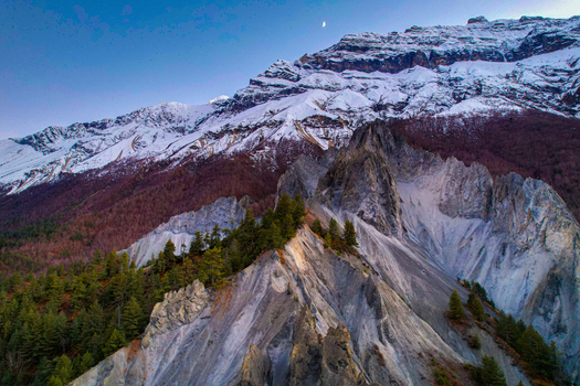 Aerial view of rugged snow capped mountain peaks rocky ridges and pine forests under a clear sky with a crescent moon in Nepal.