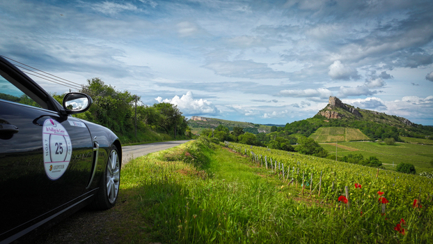 Solutre Pouilly France 01 June 2024 View of the Rock of Solutre overlooking lush green vineyards and a black car parked on a country road under a cloudy sky.