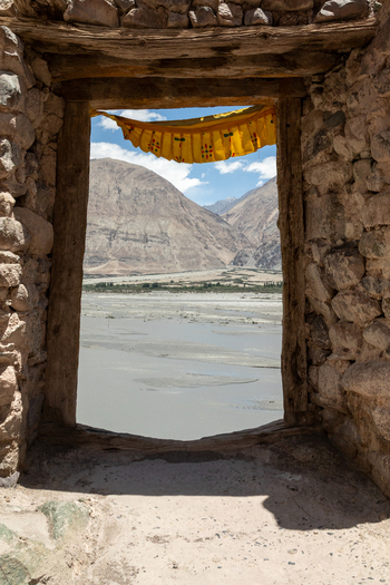 View of Charasa Gonpa framed by a rustic stone doorway with a yellow prayer flag overlooking the river valley and rugged mountains in Charasa Ladakh India.
