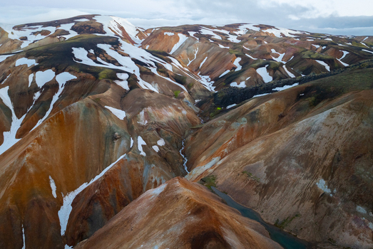 Aerial view of Landmannalaugar featuring colorful rhyolite mountains with patches of snow and a winding stream in the volcanic landscape of Rangarthing ytra Iceland. Aerial view of Landmannalaugar featuring colorful rhyolite mountains with patches of snow and a winding stream in the volcanic landscape of Rangarthing ytra Iceland.