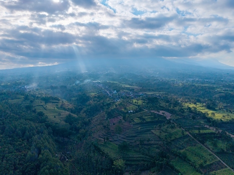 Aerial view of lush terraced fields and rolling hills in Malang with dramatic sunlight filtering through clouds over distant mountains Malang East Java Indonesia. Aerial view of lush terraced fields and rolling hills in Malang with dramatic sunlight filtering through clouds over distant mountains Malang East Java Indonesia.
