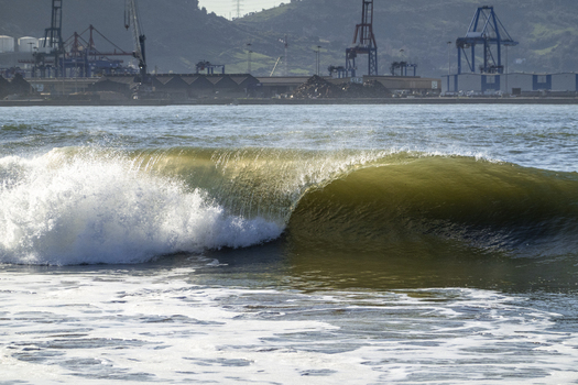 View of a powerful wave breaking at Ereaga beach with industrial port cranes and green hills in the background Algorta Euskadi Spain.