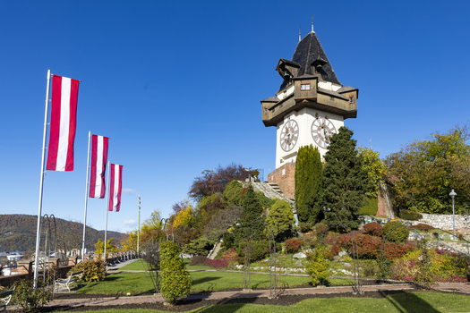View of the iconic clock tower Uhrturm with Austrian flags waving gently in the breeze amidst manicured lawns and colorful autumn foliage Graz Styria Austria. View of the iconic clock tower Uhrturm with Austrian flags waving gently in the breeze amidst manicured lawns and colorful autumn foliage Graz Styria Austria.