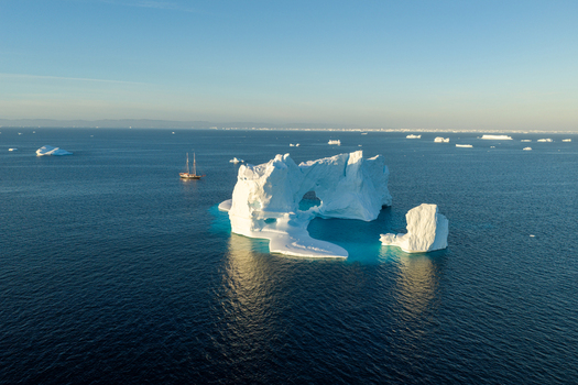 Aerial view of a large arch iceberg floating in the deep blue waters of the Arctic with a sailing ship nearby and distant icebergs under a clear sky Greenland. Aerial view of a large arch iceberg floating in the deep blue waters of the Arctic with a sailing ship nearby and distant icebergs under a clear sky Greenland.