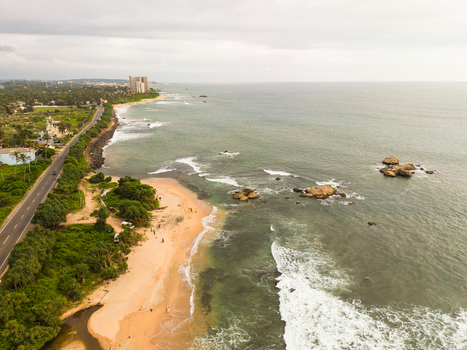 Aerial view of Dadalla beach featuring waves crashing on the sandy shore and a coastal road lined with tropical trees in Galle Southern Province Sri Lanka. Aerial view of Dadalla beach featuring waves crashing on the sandy shore and a coastal road lined with tropical trees in Galle Southern Province Sri Lanka.