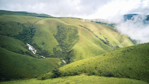 Aerial view of lush undulating green hills kissed by soft white clouds revealing a cascading waterfall amidst verdant slopes Obudu Cross River Nigeria.