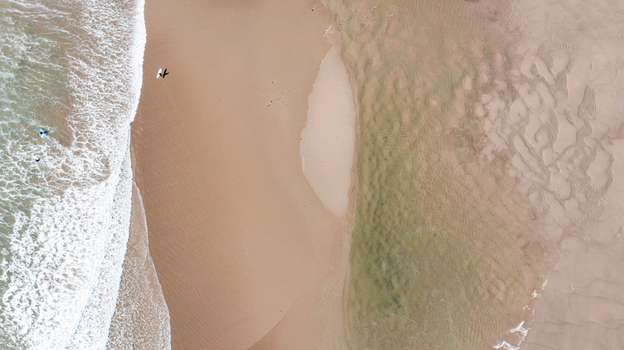 Aerial view of surfers walking on the sandy shore of Bantham Beach with crashing waves and textured sand ripples in Kingsbridge Devon United Kingdom. Aerial view of surfers walking on the sandy shore of Bantham Beach with crashing waves and textured sand ripples in Kingsbridge Devon United Kingdom.