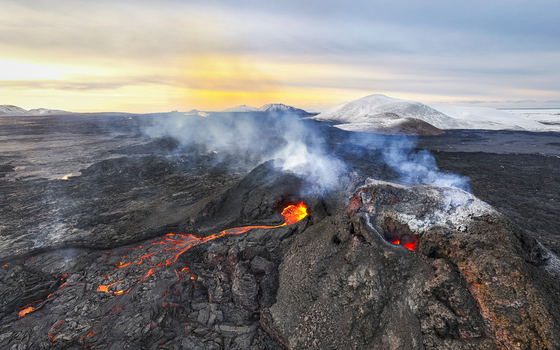 Aerial view of fiery lava flows carving paths through the dark rugged landscape near snow capped mountains under a pastel sky Reykjavik Reykjanes Iceland.