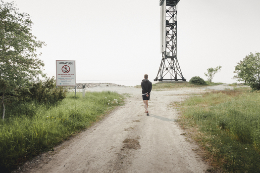 Saxby Laane maakond Estonia 01 June 2025 View of a person walking along a dirt path towards the Saxby rannik coast passing a warning sign and a tall metal beacon structure under a bright sky.
