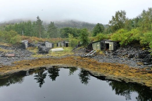 Aerial view of small wooden huts situated near a dark pool of water surrounded by rocky terrain lush trees and misty mountains in Glencoe Scotland United Kingdom.