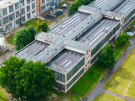 Aerial view of a large industrial building featuring solar panels on its flat roof surrounded by lush green trees and grassy areas in Olaine Olaine Municipality Latvia. Aerial view of a large industrial building featuring solar panels on its flat roof surrounded by lush green trees and grassy areas in Olaine Olaine Municipality Latvia.