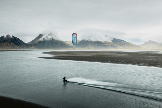 Aerial view of a kite surfer gliding across the water with the dramatic Vestrahorn mountains in the background under a cloudy sky in Sveitarfelagid Hornafjordur Iceland. Aerial view of a kite surfer gliding across the water with the dramatic Vestrahorn mountains in the background under a cloudy sky in Sveitarfelagid Hornafjordur Iceland.