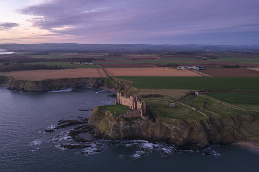 Aerial view of Tantallon Castle on a rocky cliff overlooking the sea with agricultural fields under a purple sky North Berwick Scotland United Kingdom.