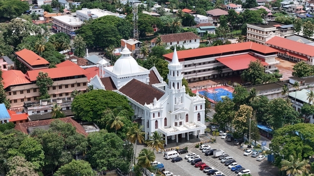 Aerial view of Mount Carmel Roman Catholic Cathedral featuring its white facade dome and spire surrounded by lush trees and a parking area in Alappuzha Kerala India. Aerial view of Mount Carmel Roman Catholic Cathedral featuring its white facade dome and spire surrounded by lush trees and a parking area in Alappuzha Kerala India.