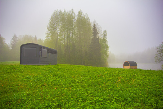 View of a modern rectangular cabin and a barrel sauna on a lush green meadow by a misty lake surrounded by trees in Latvia. View of a modern rectangular cabin and a barrel sauna on a lush green meadow by a misty lake surrounded by trees in Latvia.