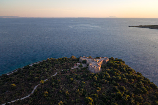 Aerial view of the triangular Porto Palermo Castle on a lush green peninsula overlooking the calm blue sea during golden hour in Porto Palermo Qarku i Vlores Albania. Aerial view of the triangular Porto Palermo Castle on a lush green peninsula overlooking the calm blue sea during golden hour in Porto Palermo Qarku i Vlores Albania.