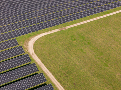 Aerial view of a large solar farm with rows of photovoltaic panels arranged in neat lines next to a dirt path and green fields in Poland.