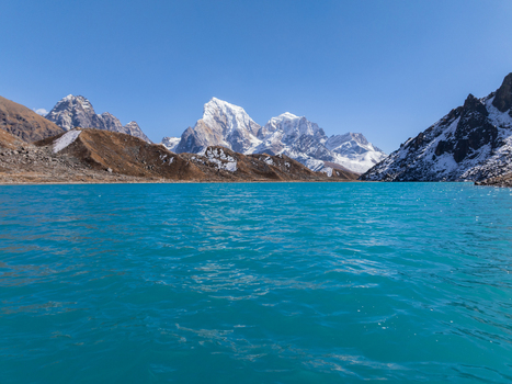 Aerial view of a vibrant turquoise glacial lake nestled beneath towering snow capped Himalayan peaks under a clear blue sky in Nepal.