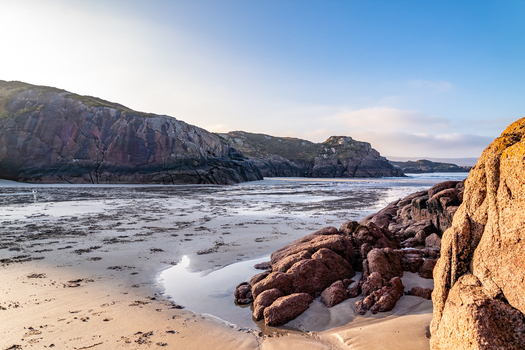 View of Cloughglass beach featuring rugged rocky cliffs golden sand and calm tidal waters under a clear blue sky in Cloughglass County Donegal Ireland. View of Cloughglass beach featuring rugged rocky cliffs golden sand and calm tidal waters under a clear blue sky in Cloughglass County Donegal Ireland.