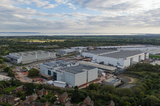 Aerial view of a large data center complex featuring industrial buildings cooling units and parking areas surrounded by green space and residential housing in the United Kingdom.