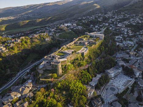 Aerial view of Gjirokaster Fortress perched on a hill overlooking the historic stone city with lush green trees and surrounding mountains in Gjirokaster Gjirokaster County Albania. Aerial view of Gjirokaster Fortress perched on a hill overlooking the historic stone city with lush green trees and surrounding mountains in Gjirokaster Gjirokaster County Albania.