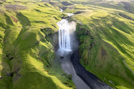 Aerial view of Skogafoss waterfall cascading down a cliff into a river surrounded by lush green hills in Rangarthing eystra Iceland.